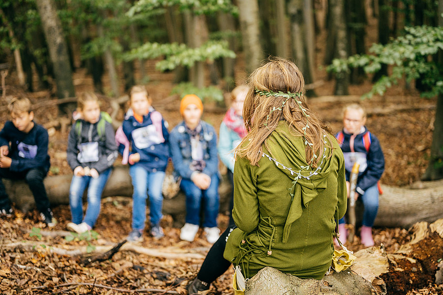 Eine Person mit langen Haaren, die mit Blättern geschmückt ist, sitzt auf einem Baumstamm und spricht. Vor ihr sitzen mehrere Kinder auf einem anderen Baumstamm und hören aufmerksam zu. Die Umgebung ist ein Wald mit Blätterteppich und Bäumen im Hintergrund.