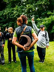 Eine Person mit einem Horn in der Hand steht mit dem Rücken zu einer Gruppe von Kindern, die ihre Hände heben. Die Umgebung besteht aus grünem Gras und Bäumen im Hintergrund.