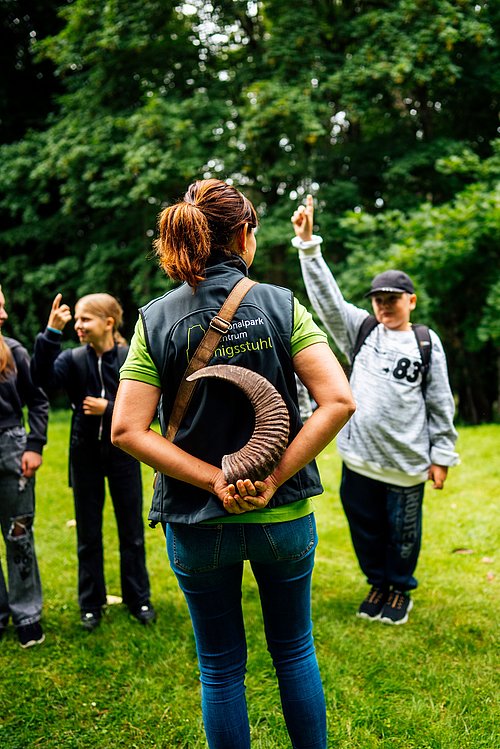 Eine Person mit einem Horn in der Hand steht mit dem Rücken zu einer Gruppe von Kindern, die ihre Hände heben. Die Umgebung besteht aus grünem Gras und Bäumen im Hintergrund.