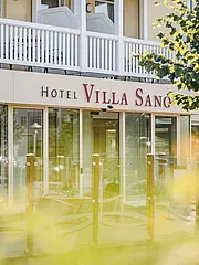 Image - Einblick in das Hotel Villa Sano. Die Fassade zeigt große Fenster und einen einladenden Eingangsbereich. Vor dem Hotel befinden sich Terrassen mit Sitzmöglichkeiten. Insgesamt vermittelt das Bild eine freundliche und einladende Atmosphäre.