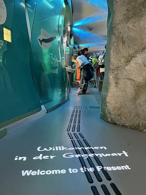 Ein moderner Ausstellungsraum mit transparenten Wänden und einem grauen Boden. Der Text „Willkommen in der Gegenwart“ und „Welcome to the present“ ist auf den Boden projiziert. Besucher erkunden die Umgebung, während im Hintergrund eine skulpturale Steinstruktur sichtbar ist.