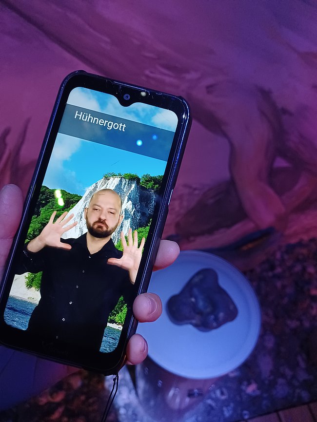 Eine Person hält ein Smartphone in der Hand. Auf dem Bildschirm ist eine digitale Darstellung mit dem Text "Hühnergott" und einer Landschaft im Hintergrund zu sehen. Der Hintergrund zeigt eine grüne Küstenlandschaft.