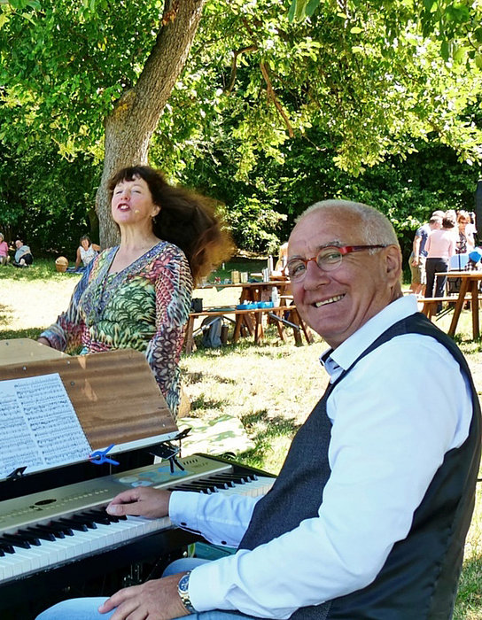 Eine fröhliche Atmosphäre im Freien, wo ein Musikant an einem Keyboard spielt, während eine Sängerin begeistert singt. Im Hintergrund sitzen und stehen Menschen auf einer Wiese, genießen ein Picknick und die Musik, umgeben von Bäumen und Sonne.