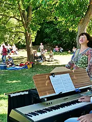 Eine fröhliche Atmosphäre im Freien, wo ein Musikant an einem Keyboard spielt, während eine Sängerin begeistert singt. Im Hintergrund sitzen und stehen Menschen auf einer Wiese, genießen ein Picknick und die Musik, umgeben von Bäumen und Sonne.