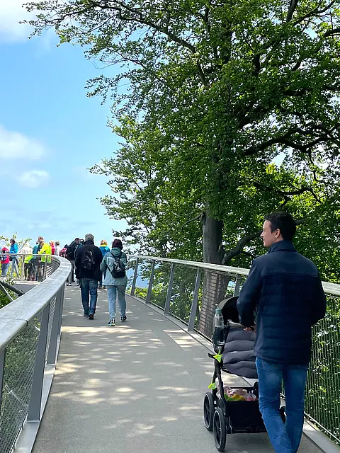 Ein schmaler Weg auf einer Aussichtsplattform, umgeben von Bäumen. Menschen gehen entlang des Weges, einige stehen an der Brüstung und genießen die Aussicht. Ein Mann schiebt einen Kinderwagen. Der Himmel ist teilweise bewölkt.