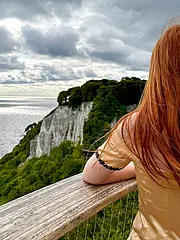 Eine person mit langen roten Haaren steht mit dem Rücken zur Kamera an einem Geländer. Im Hintergrund sind grüne Klippen und ein weites Meer unter einem bewölkten Himmel sichtbar.