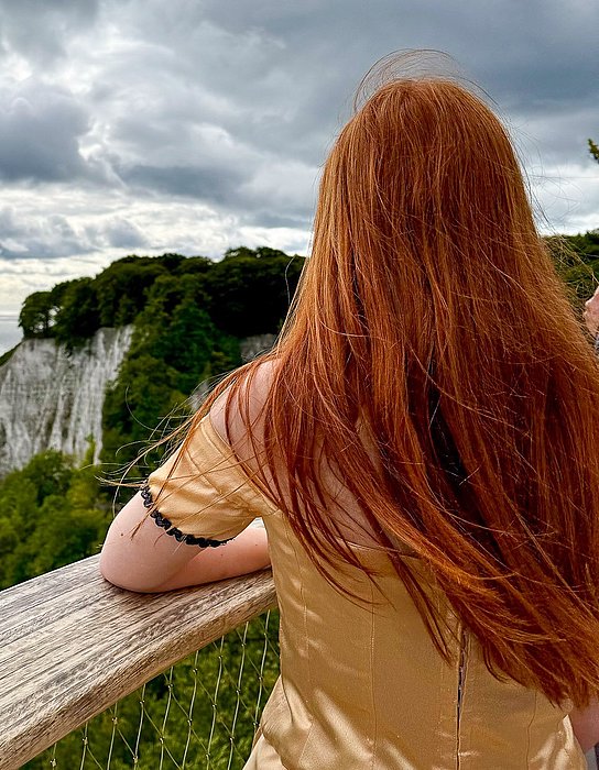 Eine person mit langen roten Haaren steht mit dem Rücken zur Kamera an einem Geländer. Im Hintergrund sind grüne Klippen und ein weites Meer unter einem bewölkten Himmel sichtbar.