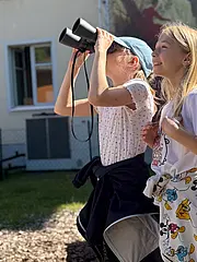 Zwei fröhliche Kinder stehen im Freien. Eines hält ein Fernglas und schaut gespannt in die Ferne, während das andere Kind begeistert lächelt. Beide tragen ungezwungene Kleidung und scheinen eine schöne Zeit zu haben. Im Hintergrund ist ein Gebäude zu erkennen.