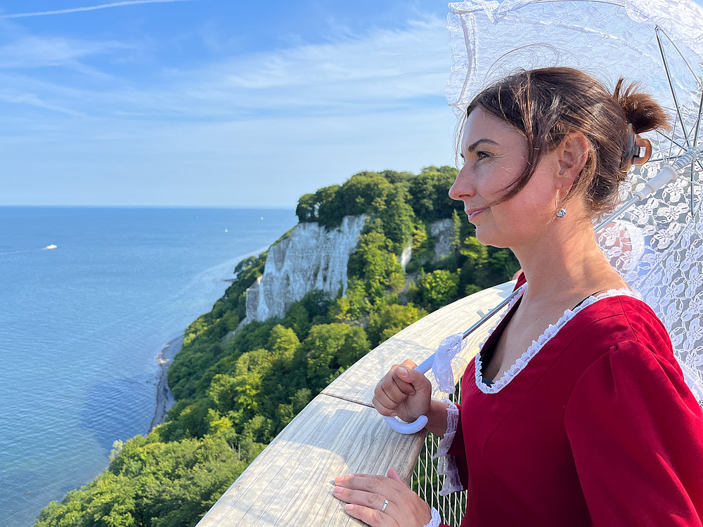 Eine Frau in einem roten Kleid steht auf einer Aussichtsplattform, hält einen weißen Stockschirm und blickt auf das Meer und die steilen Klippen im Hintergrund. Der Himmel ist blau mit wenigen Wolken, und die Umgebung ist grün bewaldet.